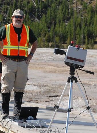 A man wearing a reflective vest and standing on barren ground stands next to a tripod bearing equipment in front of a forest